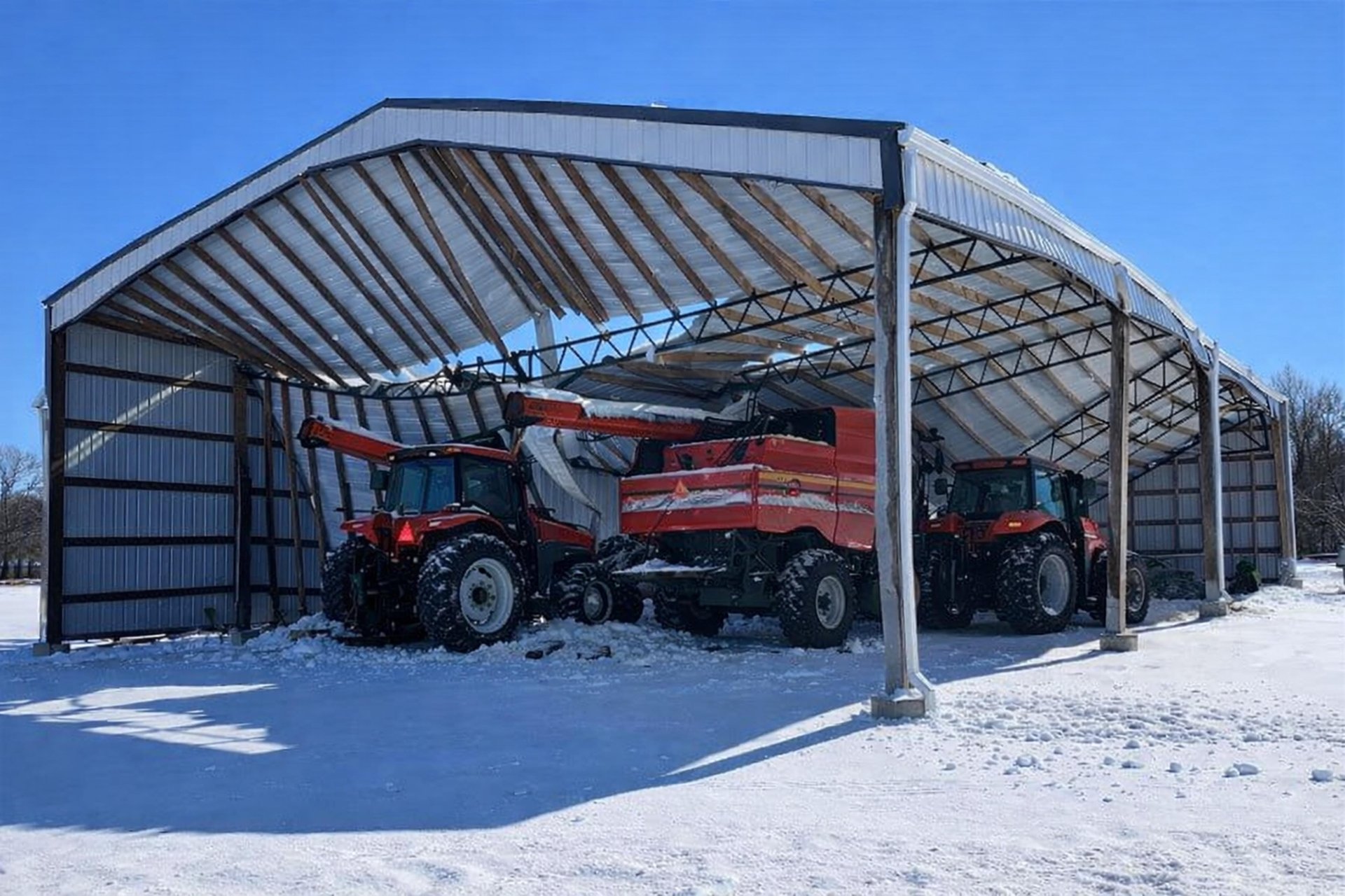 Open sided post frame equipment shed standing under a fresh snowfall with tractors parked inside